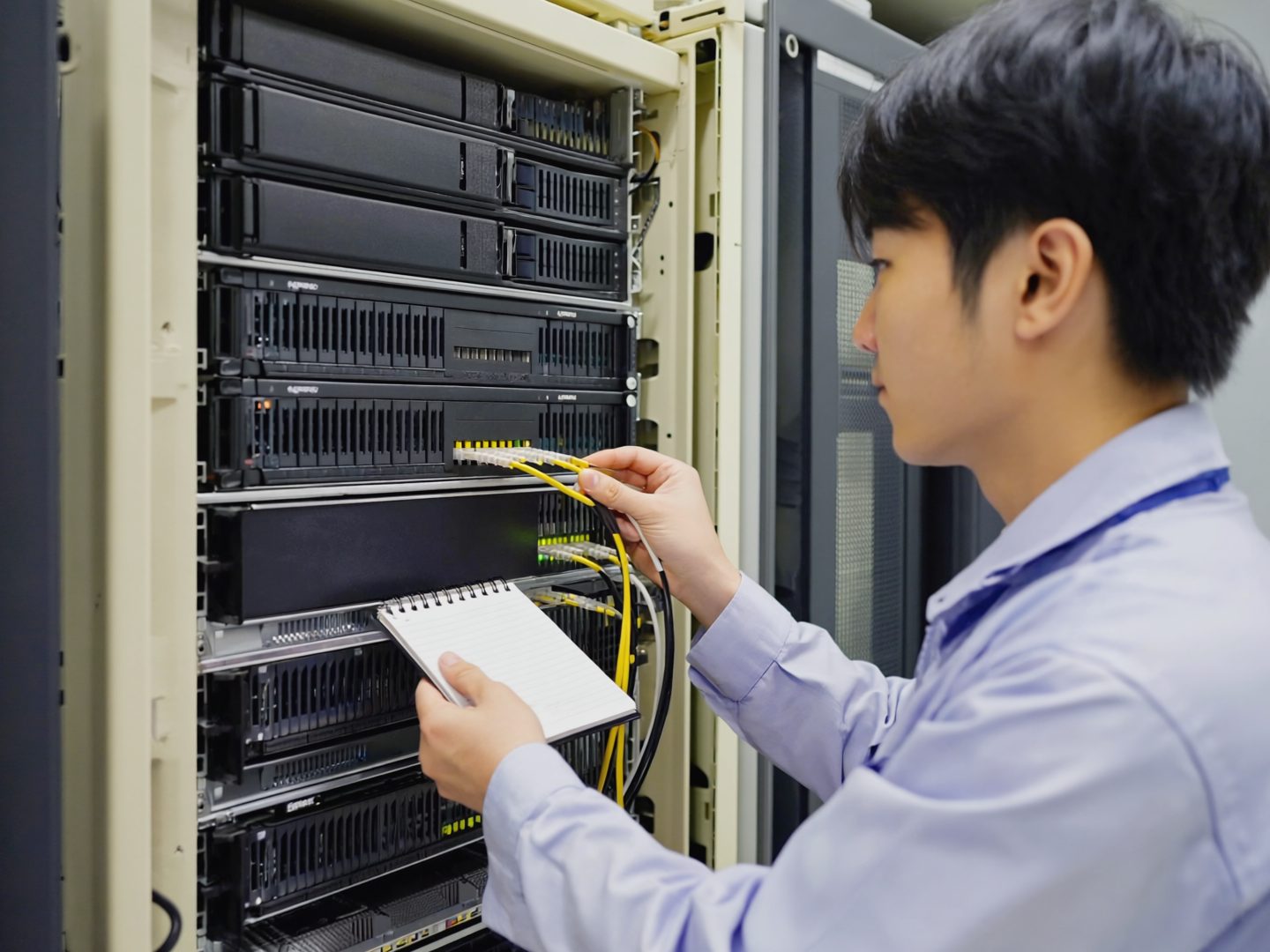 Technician connecting a device in a small server room, a notepad resting on the rack.