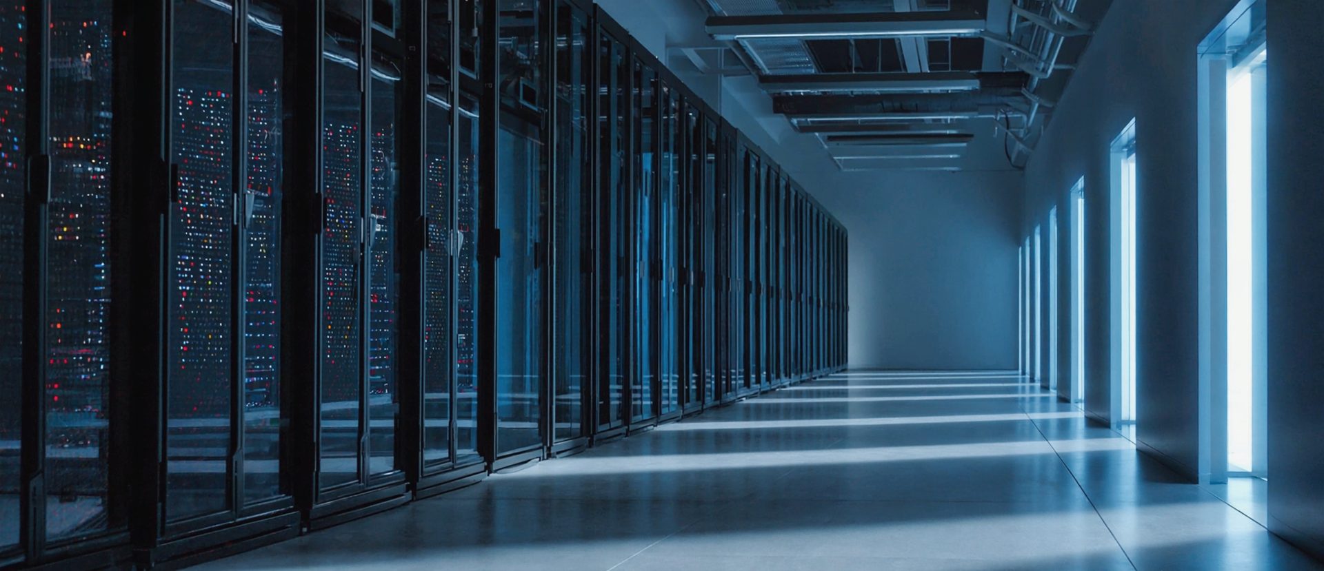Aisle between server racks in a data centre; racks with status lights on the left, daylight through narrow windows on the right.